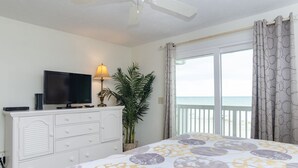 Un dormitorio con cama, televisor, lámpara, planta en maceta y balcón con vista al océano.
