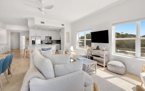 A modern living room with a sofa, coffee table, and ottoman, a dining area with chairs, a kitchen with white cabinets and appliances, and a large window offering a view. - Seagrove Beachfront Condo + Pool & Stunning Views (Santa Rosa Beach)