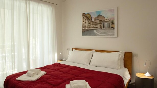 Ein Hotelzimmer mit einem großen Bett, weißer Bettwäsche, einer roten gesteppten Tagesdecke, einem Gebäude-Gemälde an der Wand, einer Nachttischlampe und einem Fenster mit durchscheinenden Vorhängen.