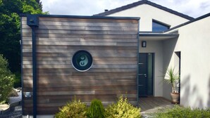 A modern building with a wooden facade, a circular window with a symbol, and a gravel driveway with small bushes. - Studio Meublé 2 Personnes (Castelmaurou)
