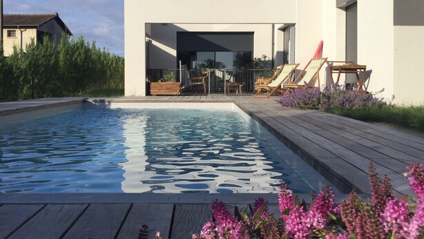 A modern outdoor pool area with a wooden deck, lounge chairs, and purple flowers in the foreground. - Studio Meublé 2 Personnes (Castelmaurou)