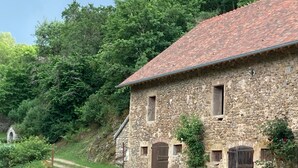A stone building with a red-tiled roof, a dirt path leading to it, and lush greenery in the background.