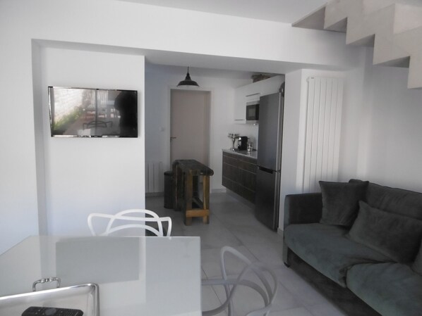 Ein modernes Wohnzimmer mit grauem Sofa, einem Essbereich mit Tisch und Stühlen sowie einer Küche mit Kühlschrank und Mikrowelle.