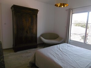 Een slaapkamer met een bed, een stoel, een kledingkast en een raam met uitzicht op gebouwen.