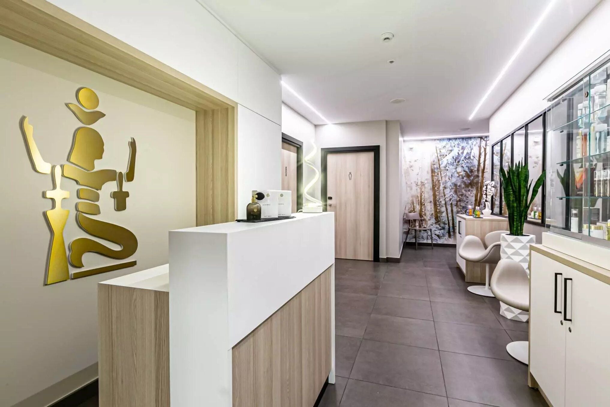 A modern reception area with a white reception desk, a wooden accent wall, and a large decorative sign. There are two doors in the background, one leading to a seating area with a plant and a vase.