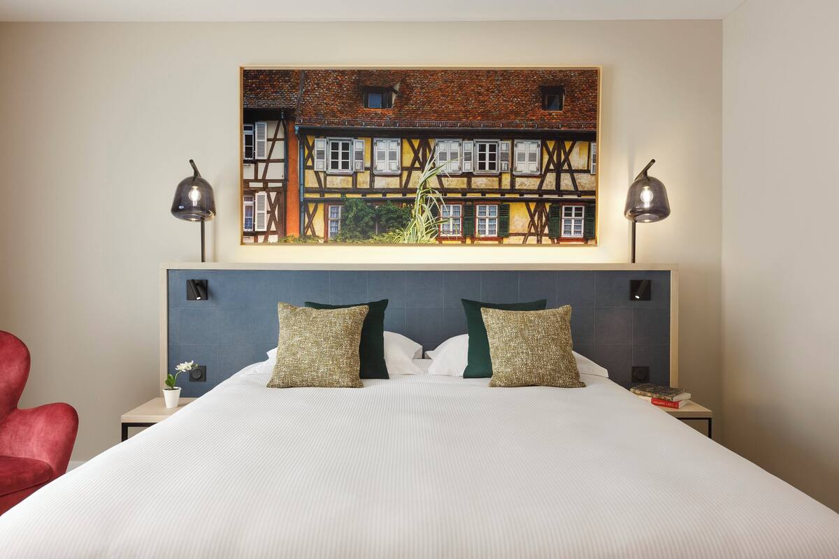 A hotel room with a large bed, two bedside lamps, a red chair, a small table with a plant, and a painting of a building on the wall.