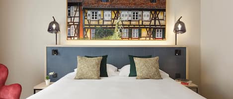 Una habitación de hotel con una cama grande, dos lámparas de noche, una silla roja, una mesita con una planta y un cuadro de un edificio en la pared.