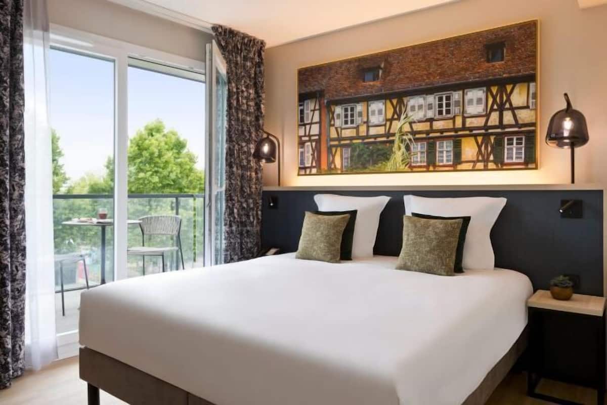 A hotel room with a large bed, a painting of a building, and a balcony with chairs.