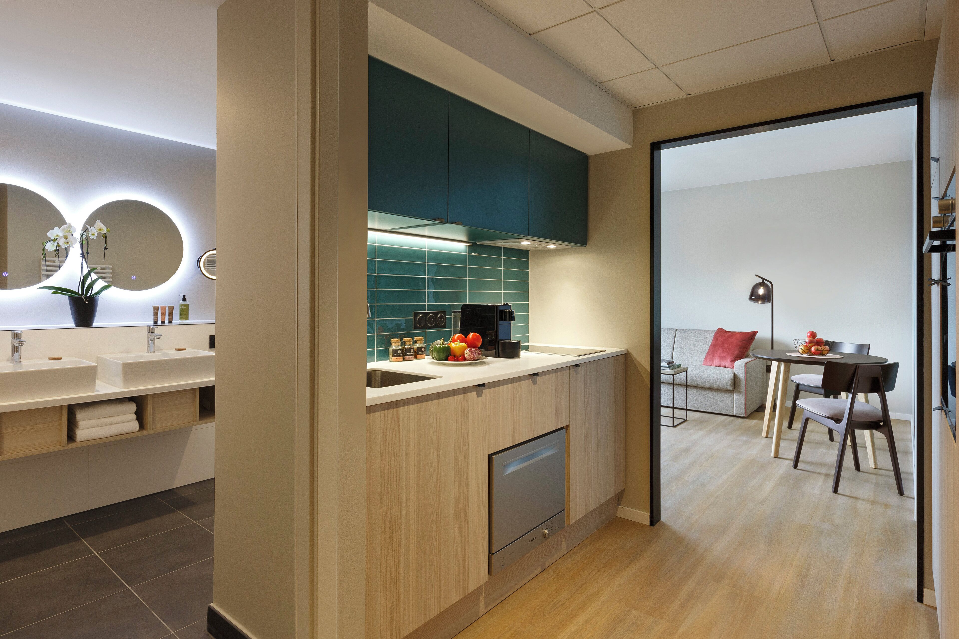 A modern kitchen with a dishwasher, a sink, and a countertop with a fruit bowl. There is a dining area with a table and chairs, and a sofa.