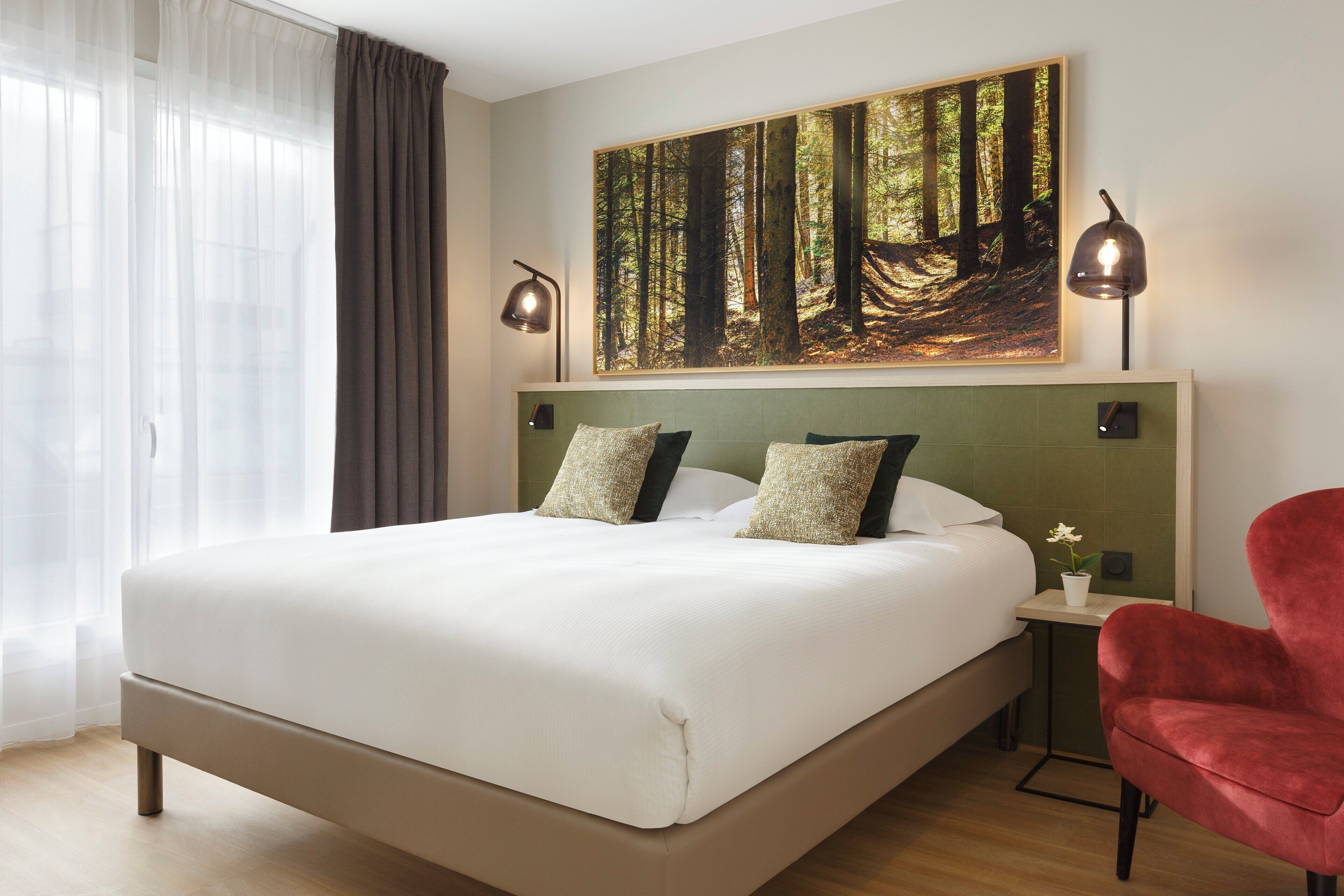 A hotel room with a large bed, two bedside lamps, a red chair, a small table with a plant, and a forest-themed painting on the wall.