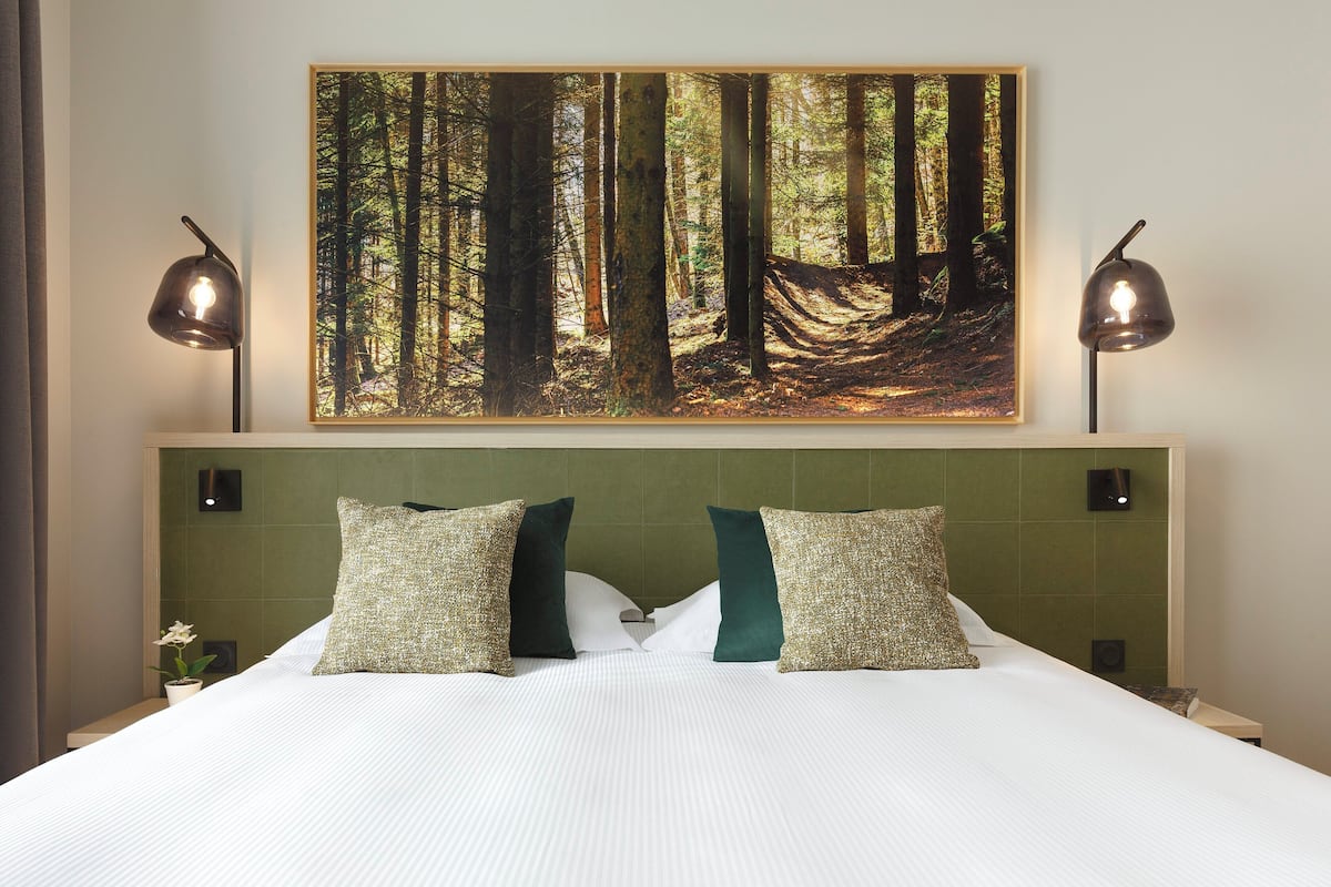 A hotel room with a large bed, two pillows, a nightstand, a wall-mounted painting of a forest, and two bedside lamps.