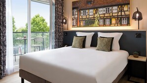Una habitación de hotel con una cama grande, dos lámparas de noche, un cuadro de un edificio y un balcón con mesa y sillas.