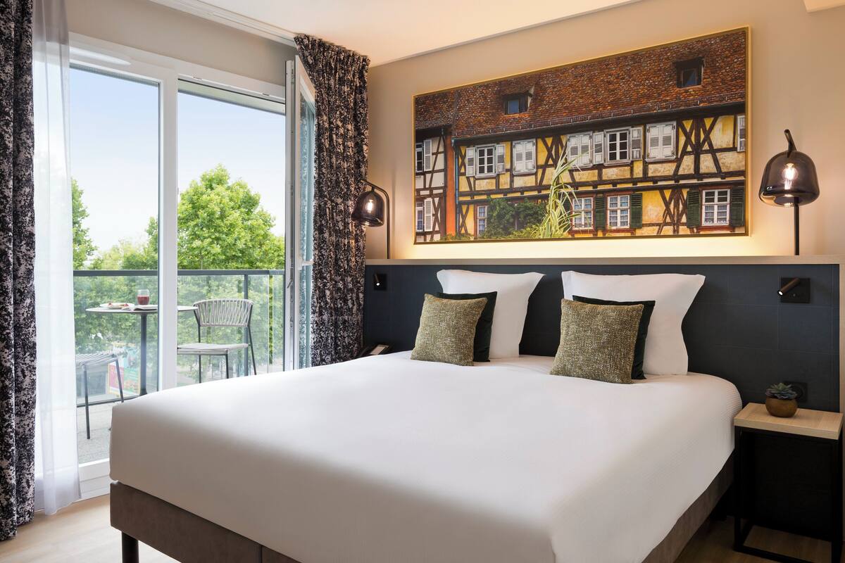 A hotel room with a large bed, two bedside lamps, a painting of a building, and a balcony with a table and chairs.