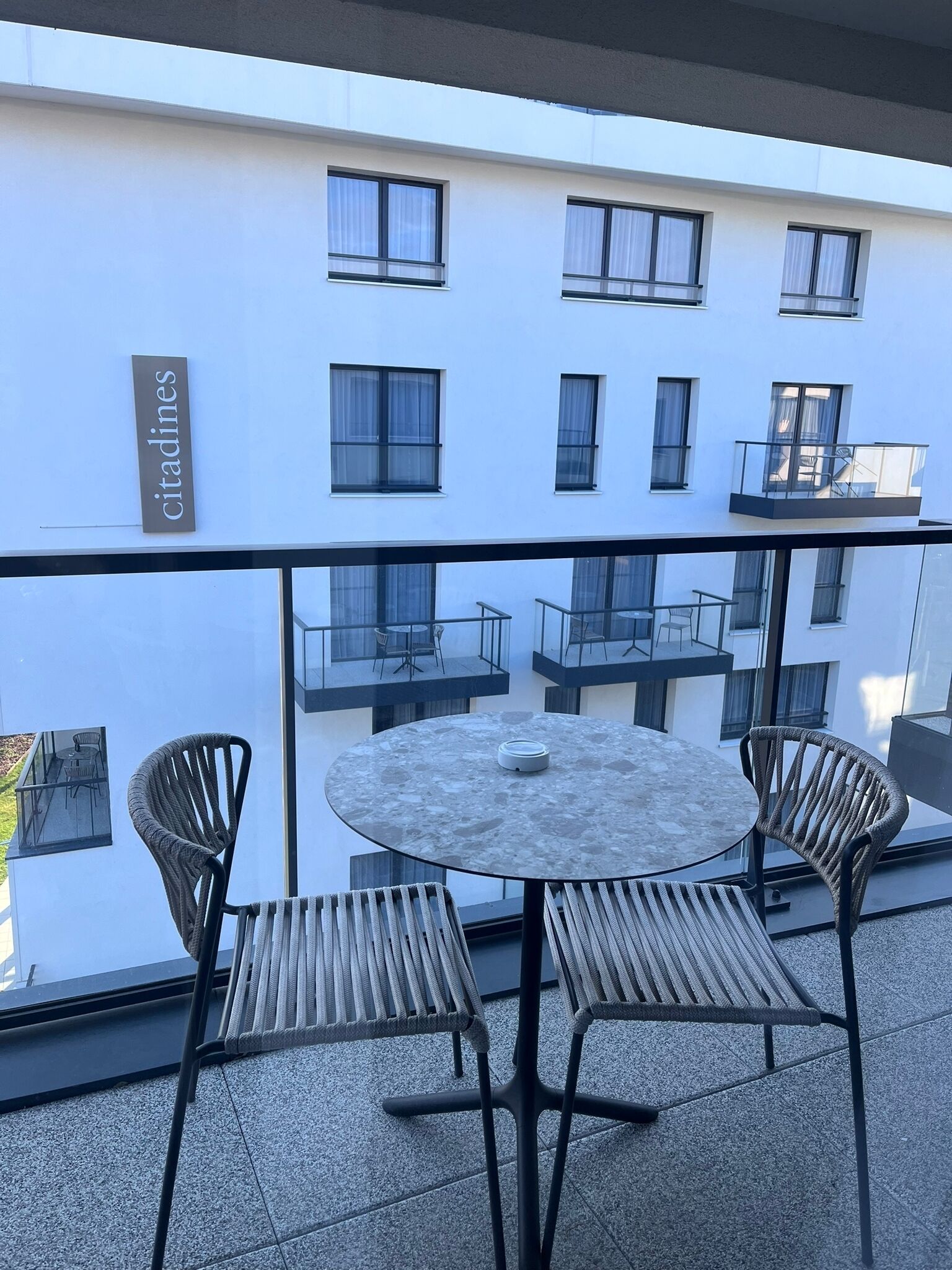 A balcony with a round table and four wicker chairs, overlooking a building with multiple windows and a sign that reads 'citadines'.