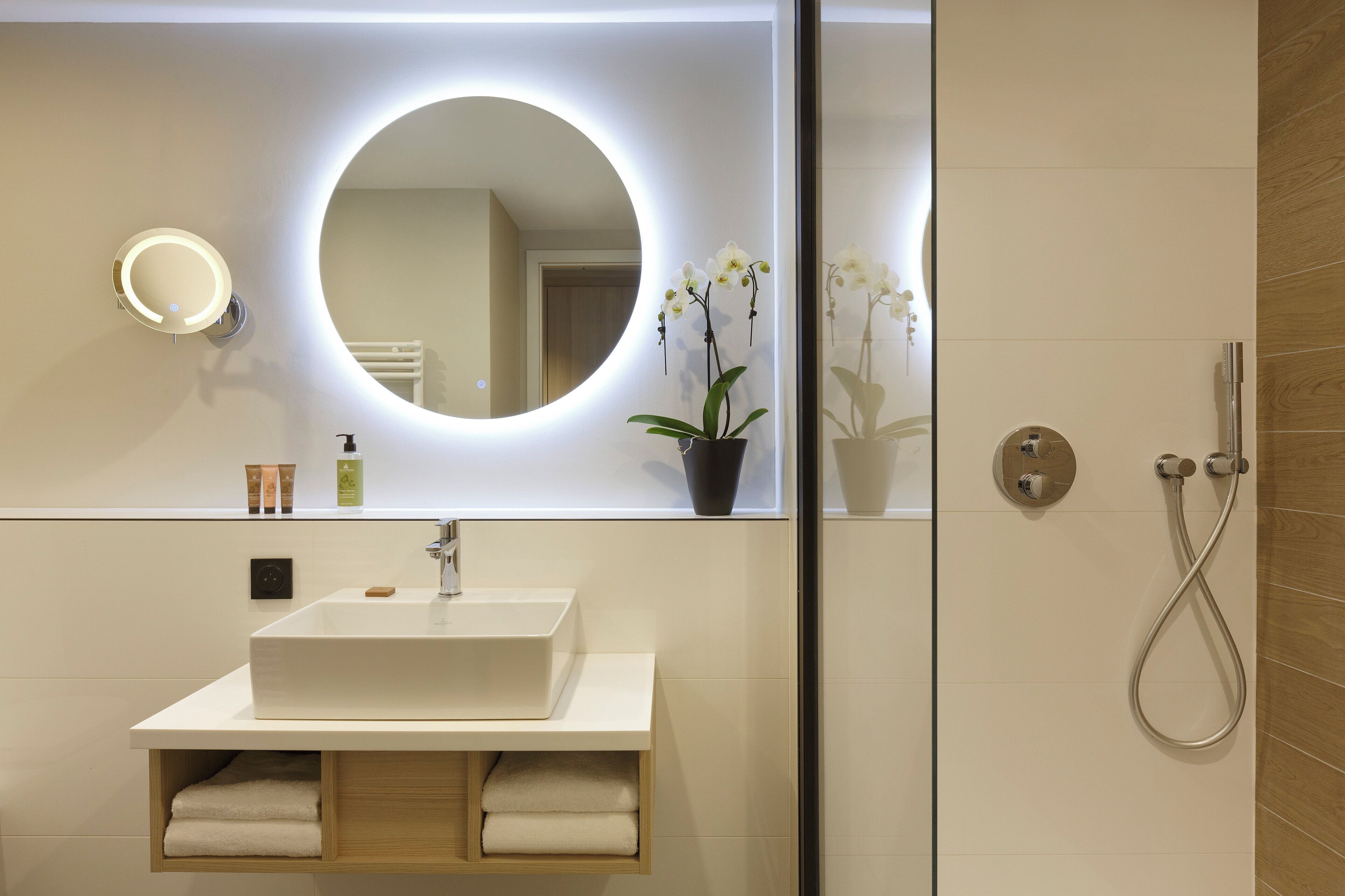 Un baño moderno con un espejo grande, un lavamanos rectangular, un cabezal de ducha de mano y una orquídea en maceta.
