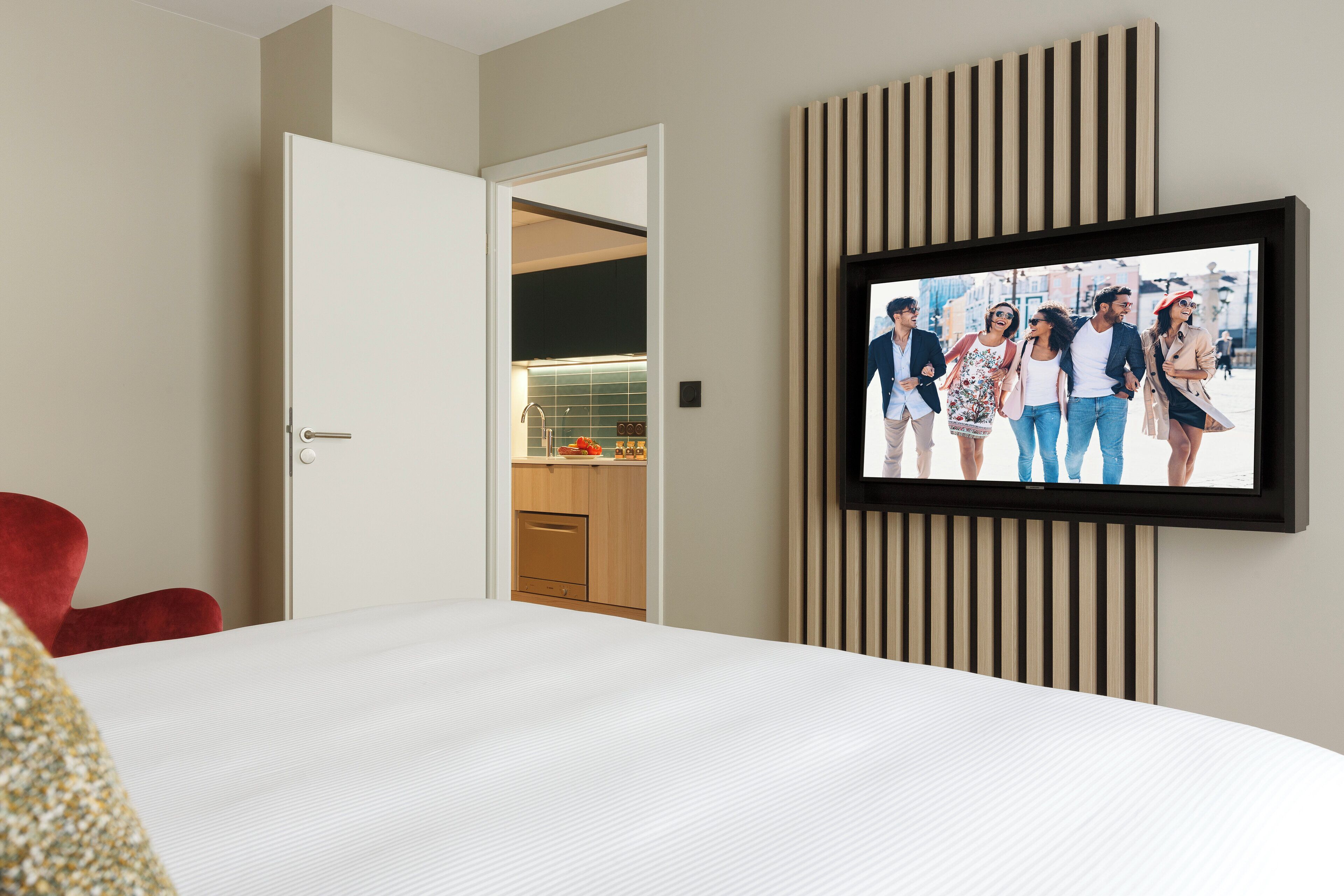 Une chambre d’hôtel avec un lit, un téléviseur fixé au mur, un fauteuil rouge et une vue sur une cuisine par une porte ouverte.