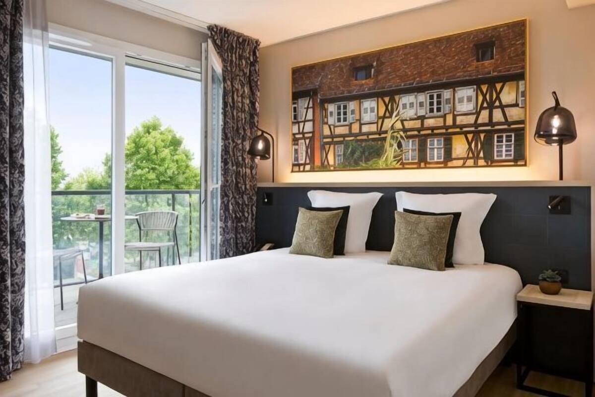 A hotel room with a large bed, a view of the outdoors, a painting on the wall, and a small table with a lamp.