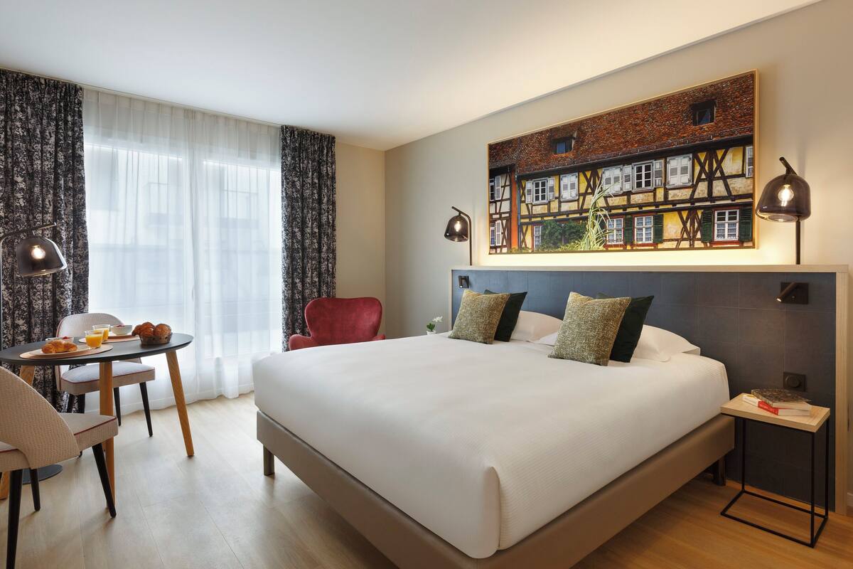 A hotel room with a large bed, a dining table with breakfast, a red chair, and a painting on the wall.