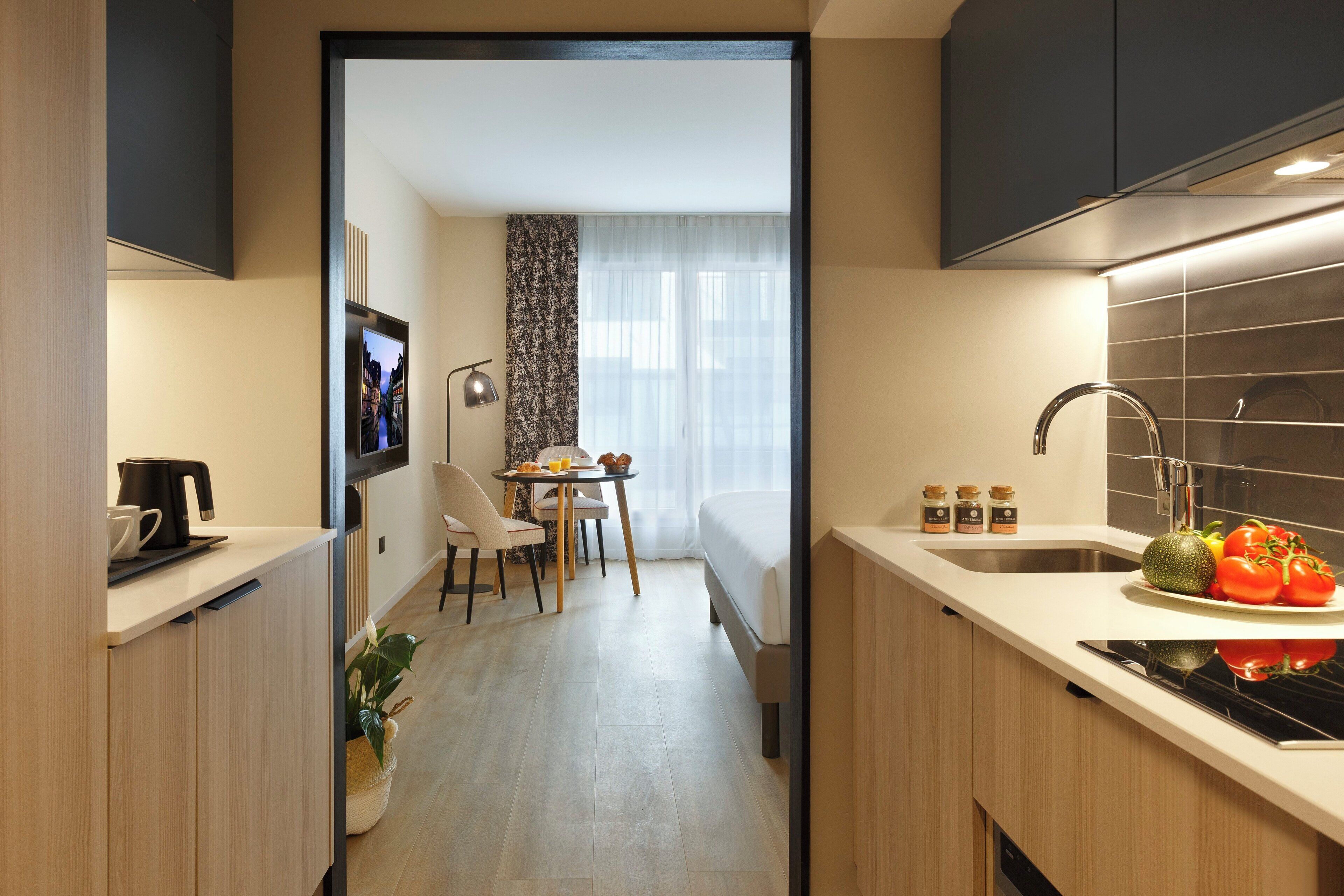 A modern kitchen with a dining area, a small table with chairs, and a bed in the background.