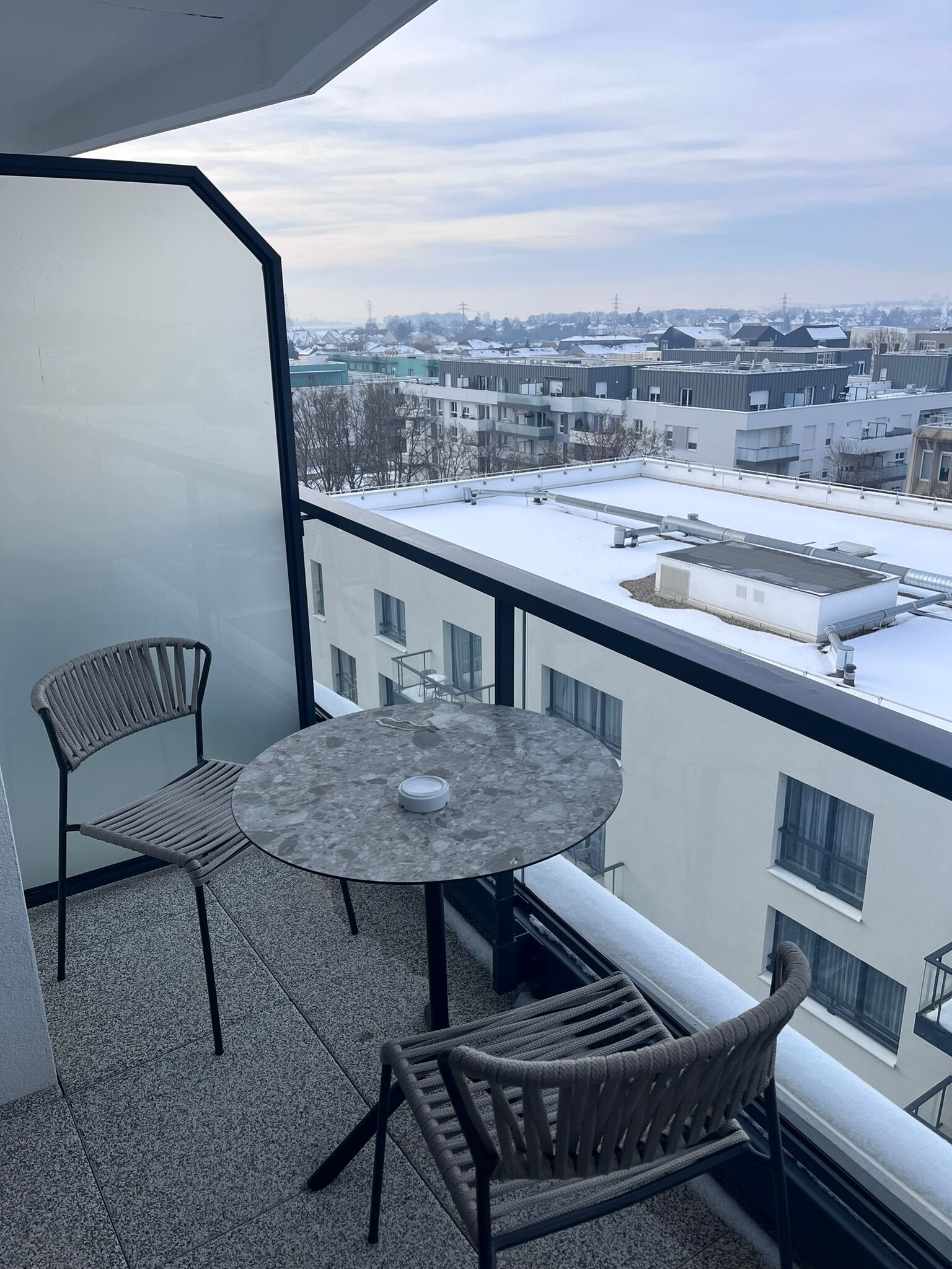 A balcony with two wicker chairs and a round table, overlooking a cityscape with buildings and a snow-covered ground.