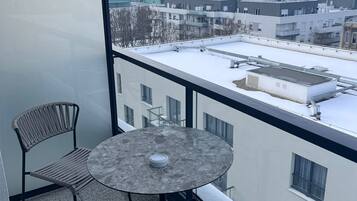 A balcony with two wicker chairs and a round table, overlooking a cityscape with buildings and a snow-covered ground.