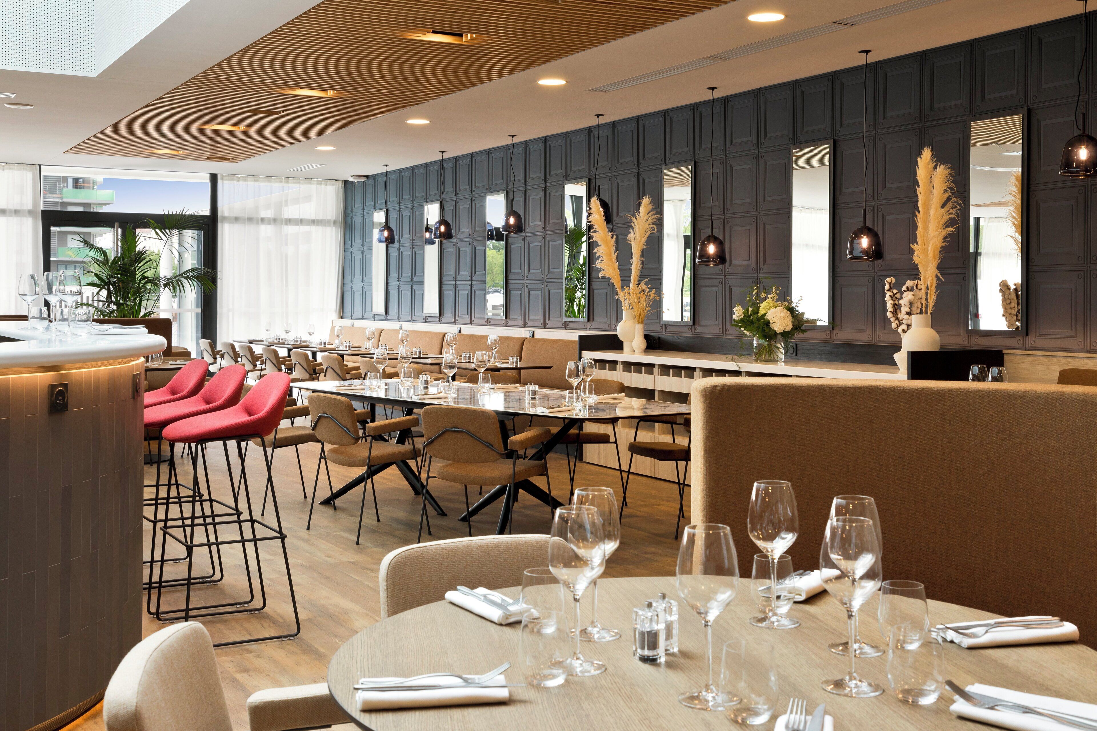 A modern restaurant with a long dining area, multiple tables, and red bar stools.