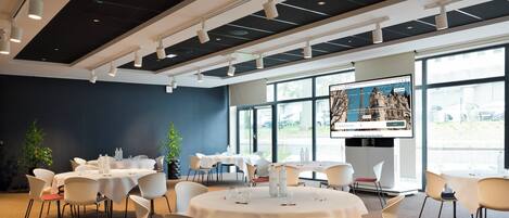 A modern dining area with round tables and chairs, a large window with a view of parked cars, and a screen displaying a map.