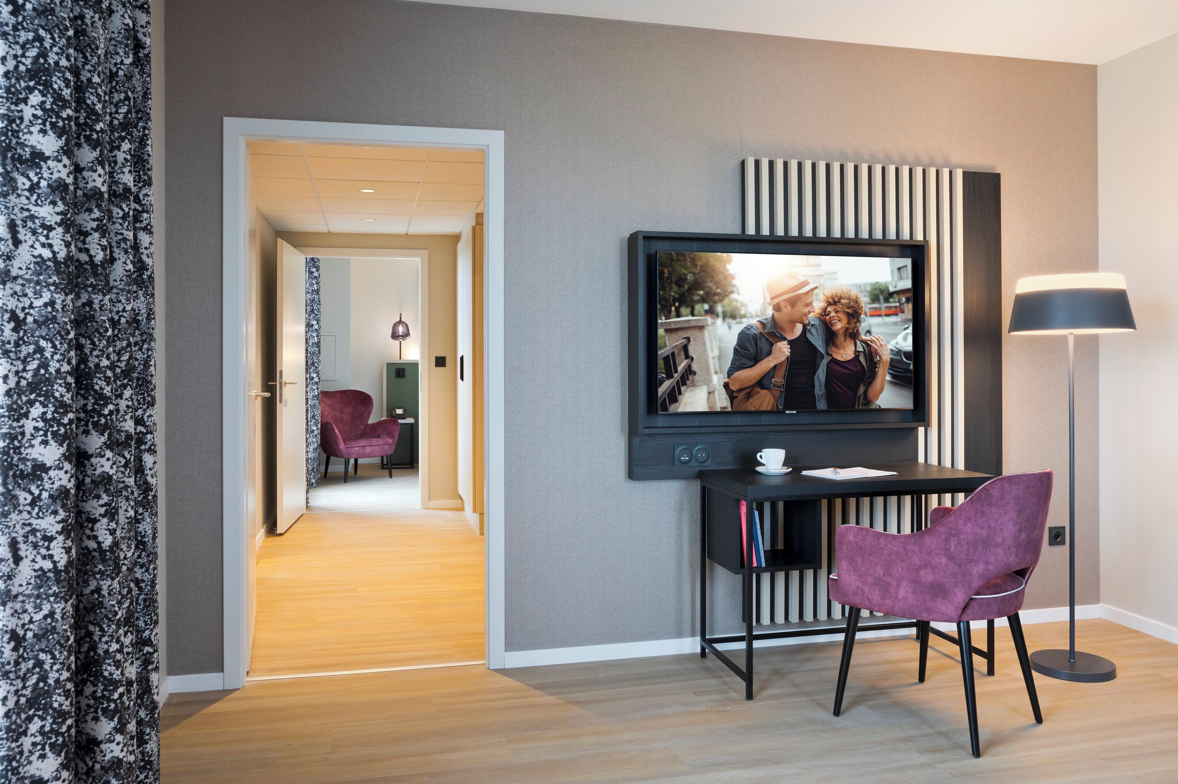 Una habitación de hotel moderna con un televisor montado en la pared, una mesita con una silla y una lámpara.