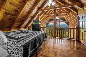 A wooden cabin with a high ceiling, a bed with a patterned blanket, a television, and a balcony with a view of the mountains. - Private, Romantic Cabin with Breathtaking Views Throughout ] Hot Tub (Sevierville)