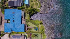 En fastighet vid kusten med ett hus som har blått tak, solpaneler, en swimmingpool och en klippig strand.