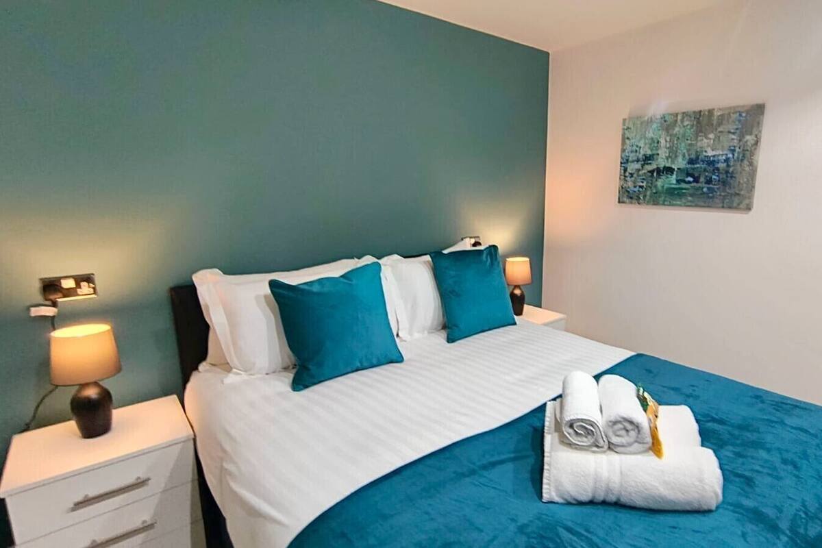 A neatly made bed with white and teal bedding, two teal pillows, and a pair of rolled towels on the bed.