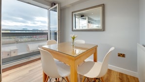 A dining area with a glass table, white chairs, a vase with flowers, and a large window with a city view. - Swan Apartments - By Week2Week (Newcastle-upon-Tyne)