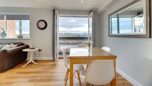 A modern living room with a glass dining table, white chairs, a brown leather sofa, a round clock, and a large window offering a city view. - Swan Apartments - By Week2Week (Newcastle-upon-Tyne)