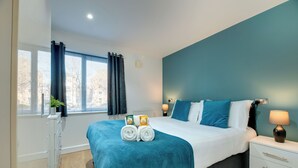 A modern bedroom with a large bed, a nightstand, a dresser, and a window with curtains. - Swan Apartments - By Week2Week (Newcastle-upon-Tyne)