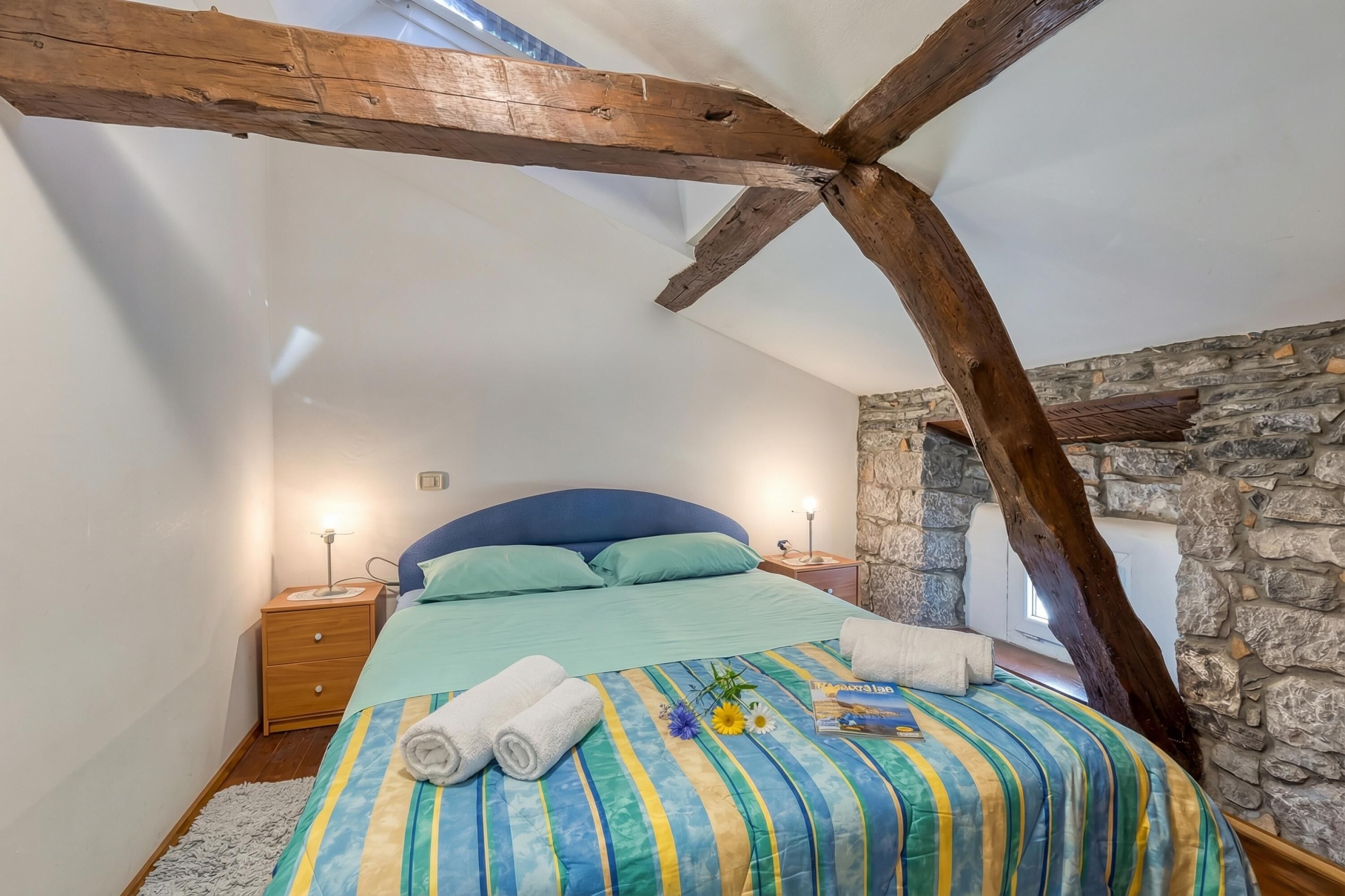 Een slaapkamer met een schuine plafond, een bed met een gestreept dekbedovertrek, twee bedlampen en een stenen muur.