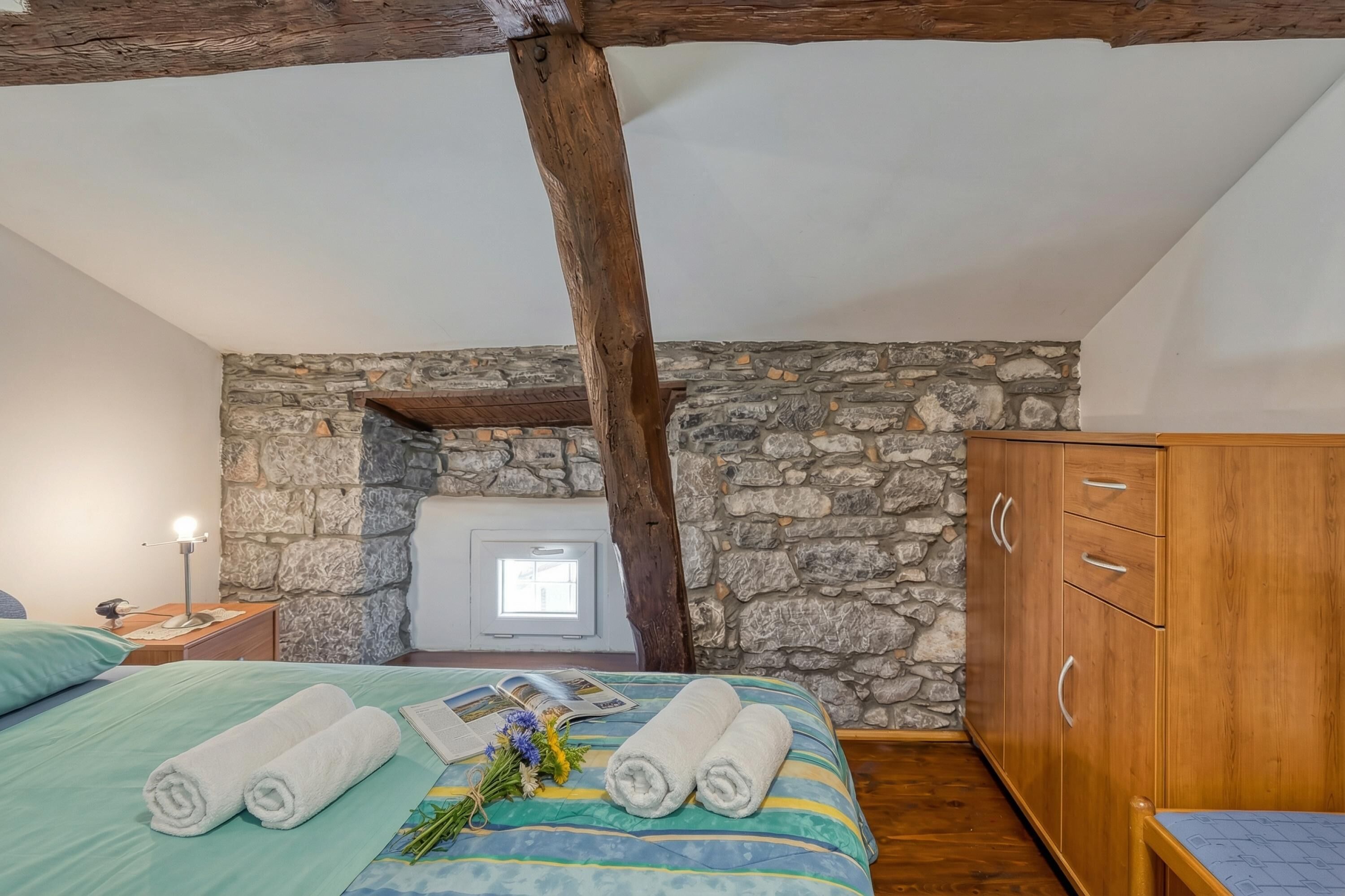 Ein Schlafzimmer mit Steinwand, Holzbalken, einem Bett mit aufgerollten Handtüchern, einem Nachttisch und einem Kleiderschrank.