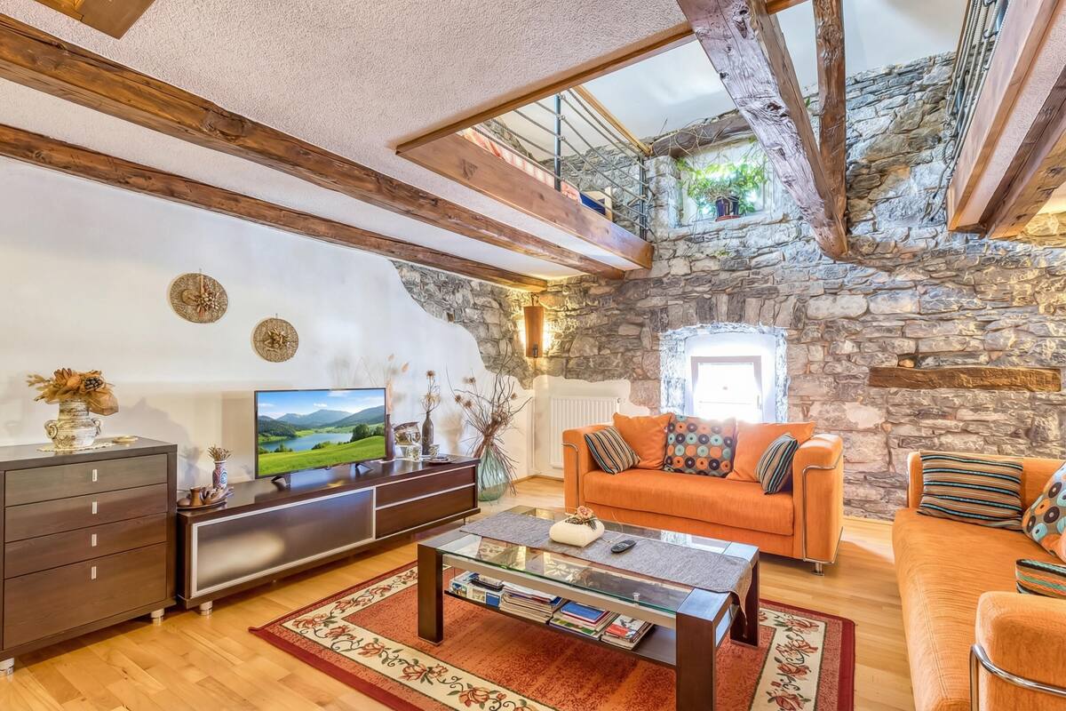 Ein Wohnzimmer mit Steinwand, Holzbalken, einer Couch, einem Couchtisch und einem Fernseher.
