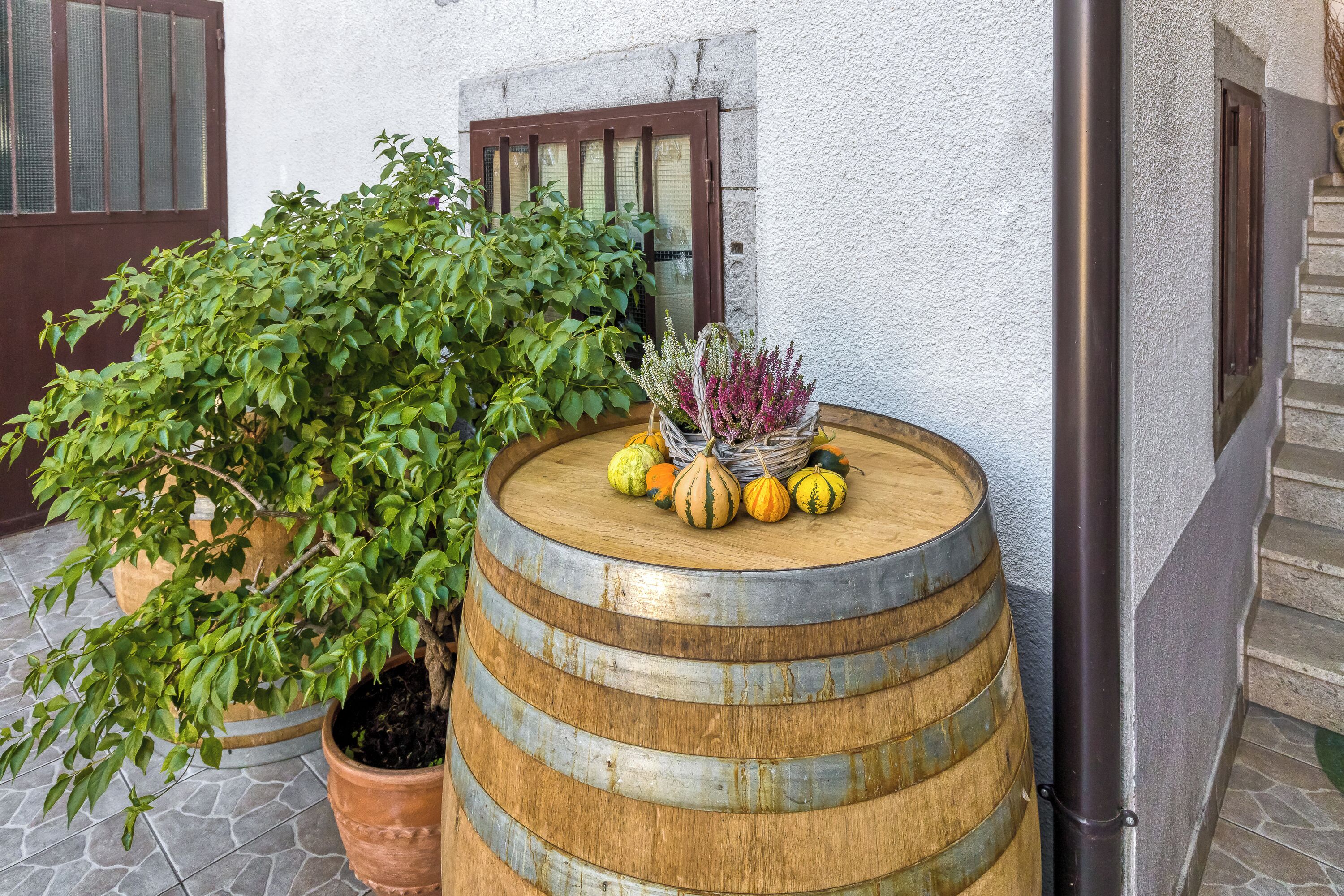 A rustic outdoor setting with a wooden barrel, a potted plant, and a wicker basket with pumpkins and flowers.
