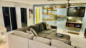 A living room with a sectional sofa, a flat-screen TV, wooden shelves with plants, and a ceiling fan. - Family Home w/Pool , On Golf Course & Near Beach !  (Fort Myers)