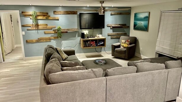 A modern living room with a sectional sofa, a television, wooden wall decor, and a ceiling fan. - Family Home w/Pool , On Golf Course & Near Beach !  (Fort Myers)