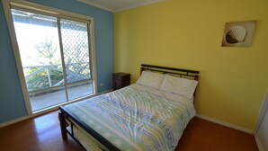 A bedroom with a bed, a small nightstand, a window with a view, and a wall-mounted fan.
