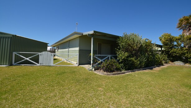 A single-story building with a flat roof and a covered porch. There is a fenced area with a gate to the left, and a small garden area with shrubs and a tree to the right.