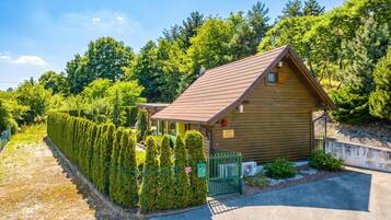 A wooden cabin with a sloped roof, surrounded by a neatly trimmed hedge and a gravel path leading to the entrance.