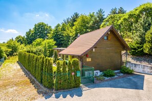 A wooden cabin with a sloped roof, surrounded by a neatly trimmed hedge and a gravel path leading to the entrance.