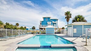 A swimming pool with a blue house in the background, surrounded by a white fence and palm trees. - Steps to Beach | Pet Friendly | Community Pools and Pier (Gulf Shores)