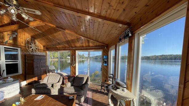 Una cabaña de madera con un gran ventanal con vistas a un lago, amueblada con sillas estampadas, una mesa de madera y una guitarra.