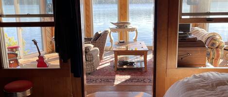 A wooden cabin room with a bed, a table, and a view of a lake.