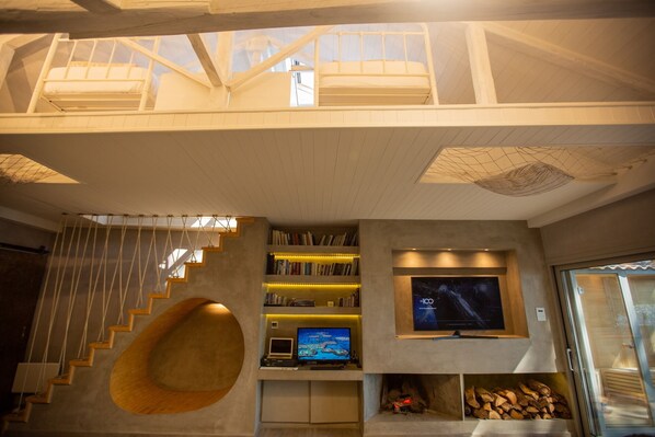 Un interior moderno con un dormitorio tipo loft, una escalera de diseño de madera, una estantería, un televisor de pantalla plana y una chimenea.