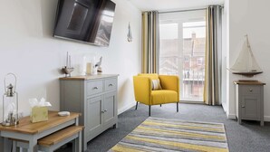 Une chambre d’hôtel moderne avec un canapé gris, une table en bois avec une lampe, un téléviseur fixé au mur et une vue sur la ville à travers de grandes fenêtres.