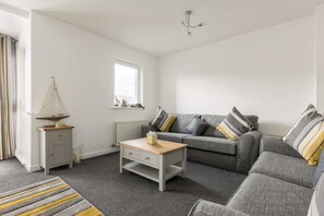 Een woonkamer met een grijze bank, een klein houten bijzettafeltje, een grijze kast, een gestreept vloerkleed en een raam met jaloezieën.